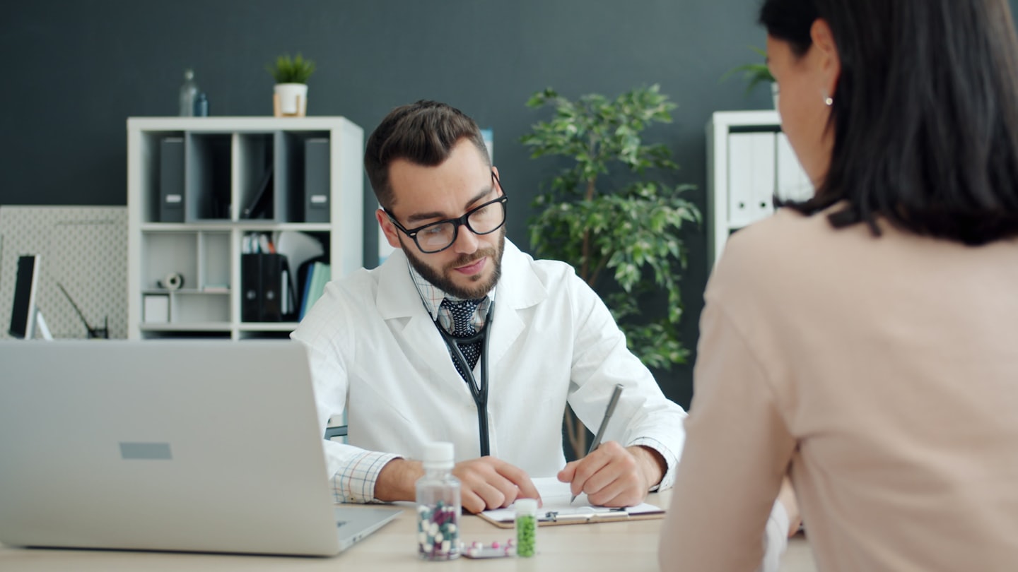 Médico escribe en portapapeles durante consulta con paciente en escritorio.
